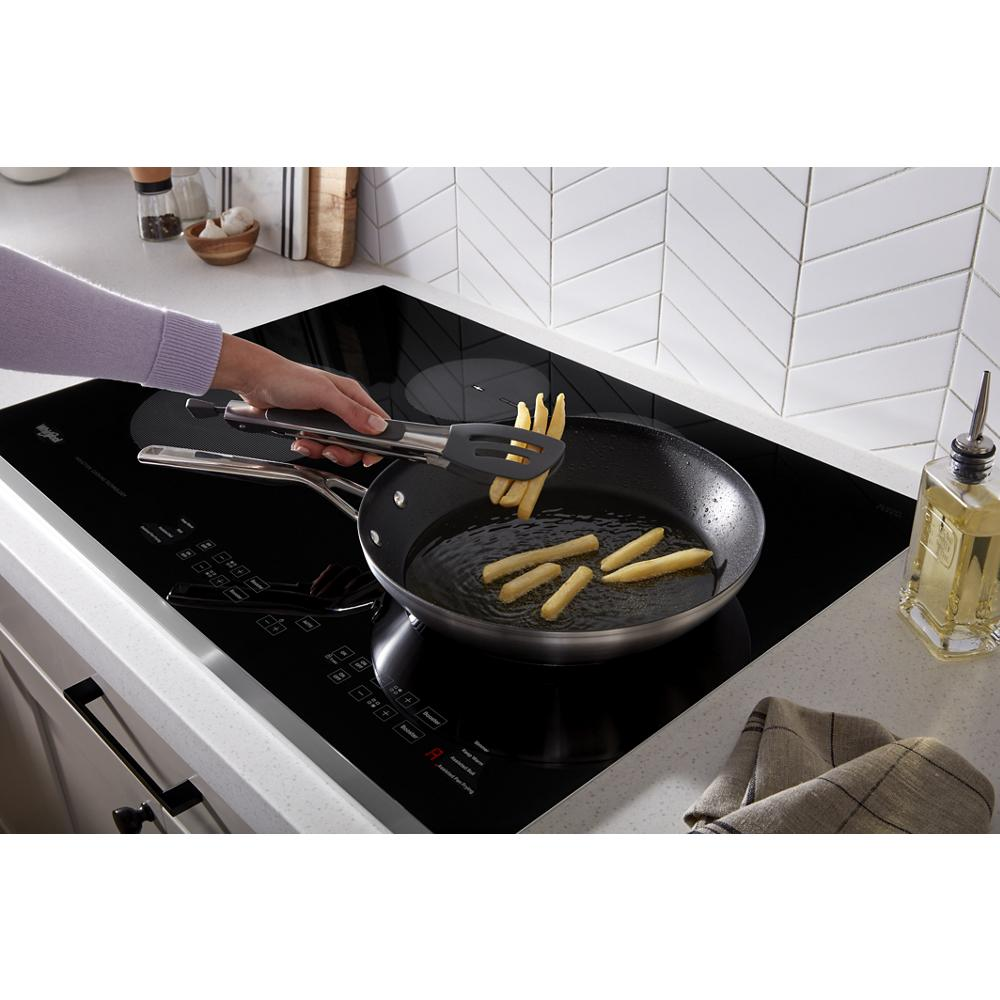 Hand using tongs to cook French fries in a black skillet on a black glass 30-inch induction cooktop in modern kitchen