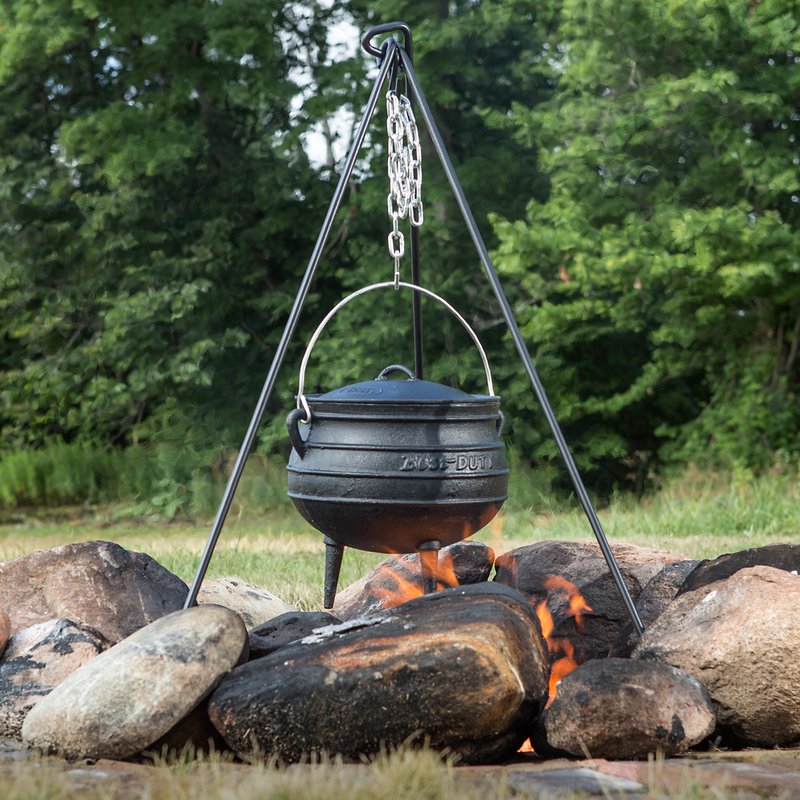 Steel tripod with adjustable chain suspending a cast iron pot over an open campfire surrounded by stones outdoors