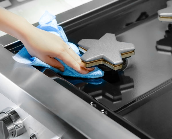 hand wiping stainless steel gas range burner with blue microfiber cloth for kitchen maintenance