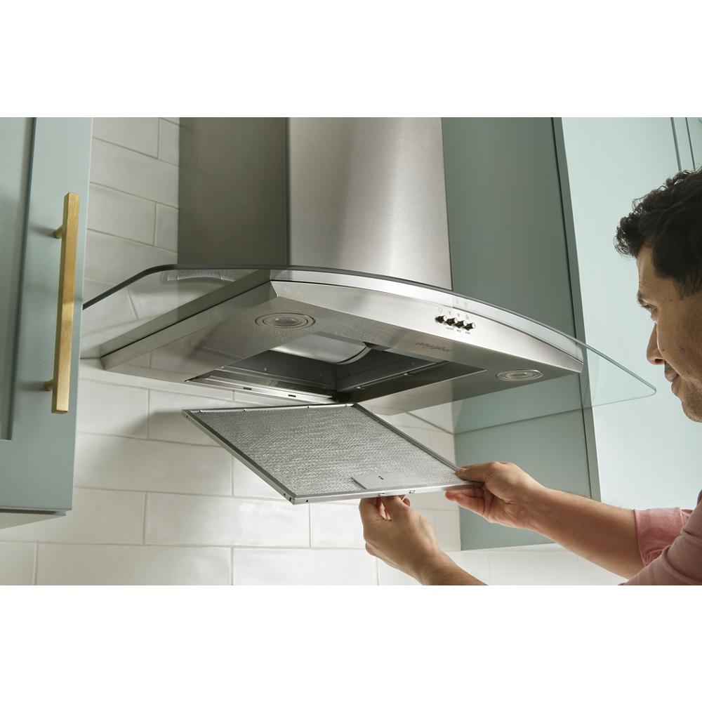 Person removing detachable metal grease filter from stainless steel curved glass wall mount range hood in kitchen appliances setting