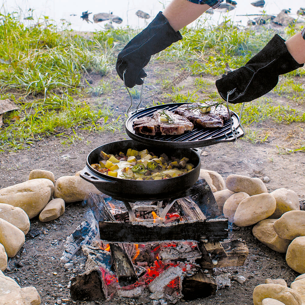 Outdoor cooking scene with cast iron grill lifting steaks above a skillet over open campfire in nature setting