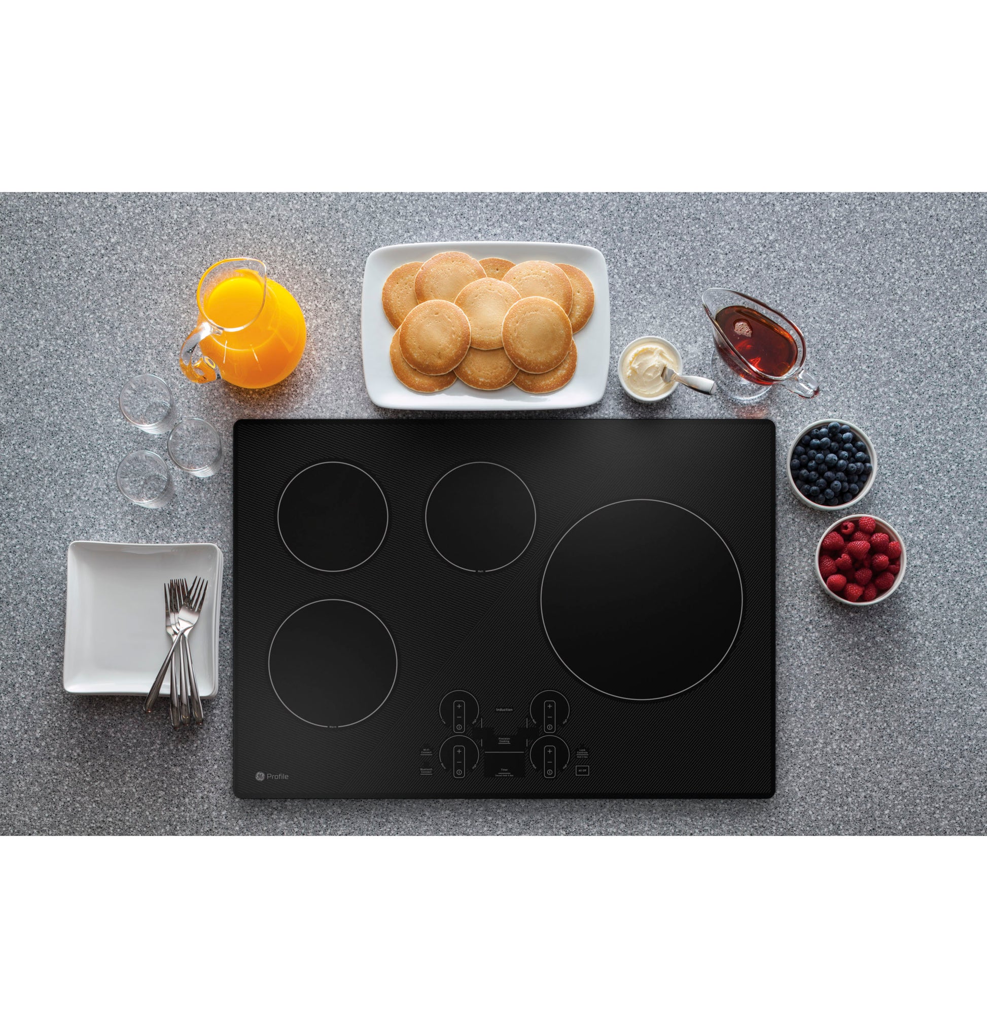 Black induction cooktop with touch controls surrounded by breakfast items including pancakes, syrup, berries, and orange juice on countertop