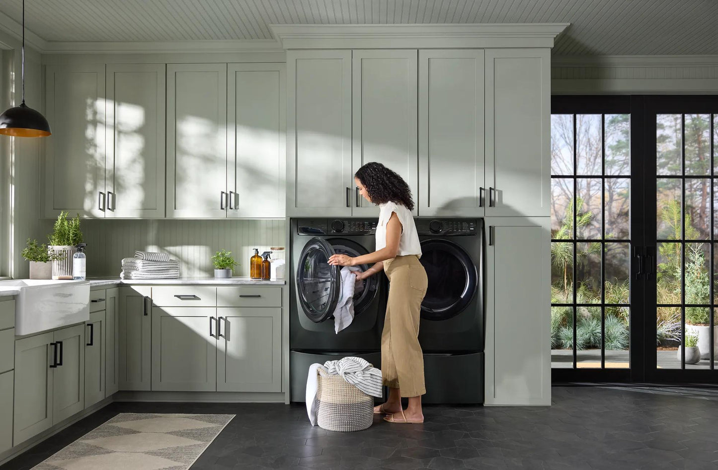 Woman loading clothes into a black front load electric dryer in a modern laundry room with gray cabinetry and natural light