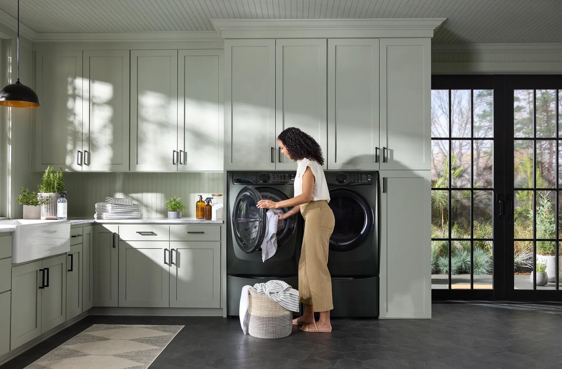 Woman loading clothes into a black front load electric dryer in a modern laundry room with gray cabinetry and natural light
