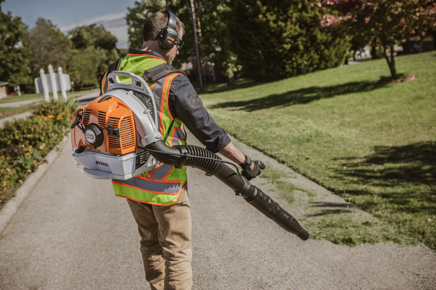 Person using orange and gray professional backpack blower on paved path wearing safety vest and ear protection for home improvement tasks