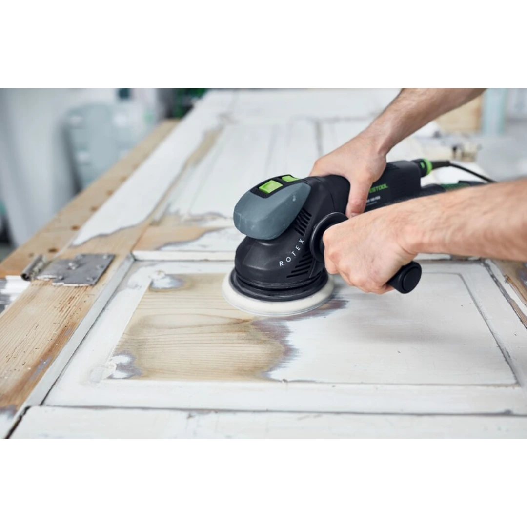 Person using a black and green handheld sander with auxiliary side handle on weathered wooden door in woodworking tool setting