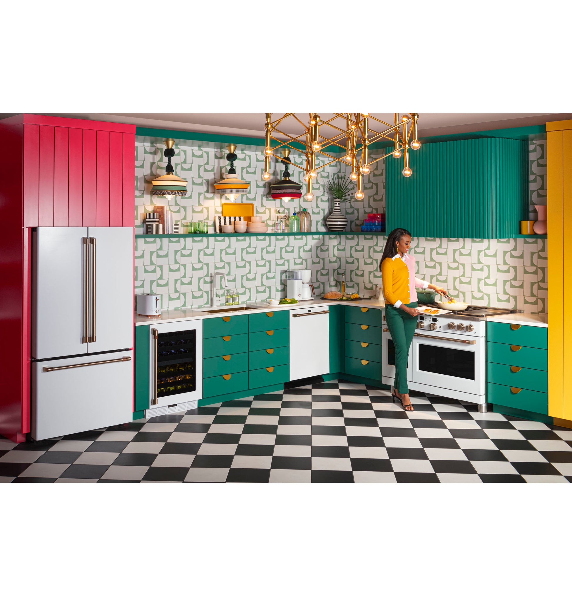 Woman cooking in colorful kitchen with teal and yellow cabinets, checkered floor, stainless appliances, and modern lighting fixture