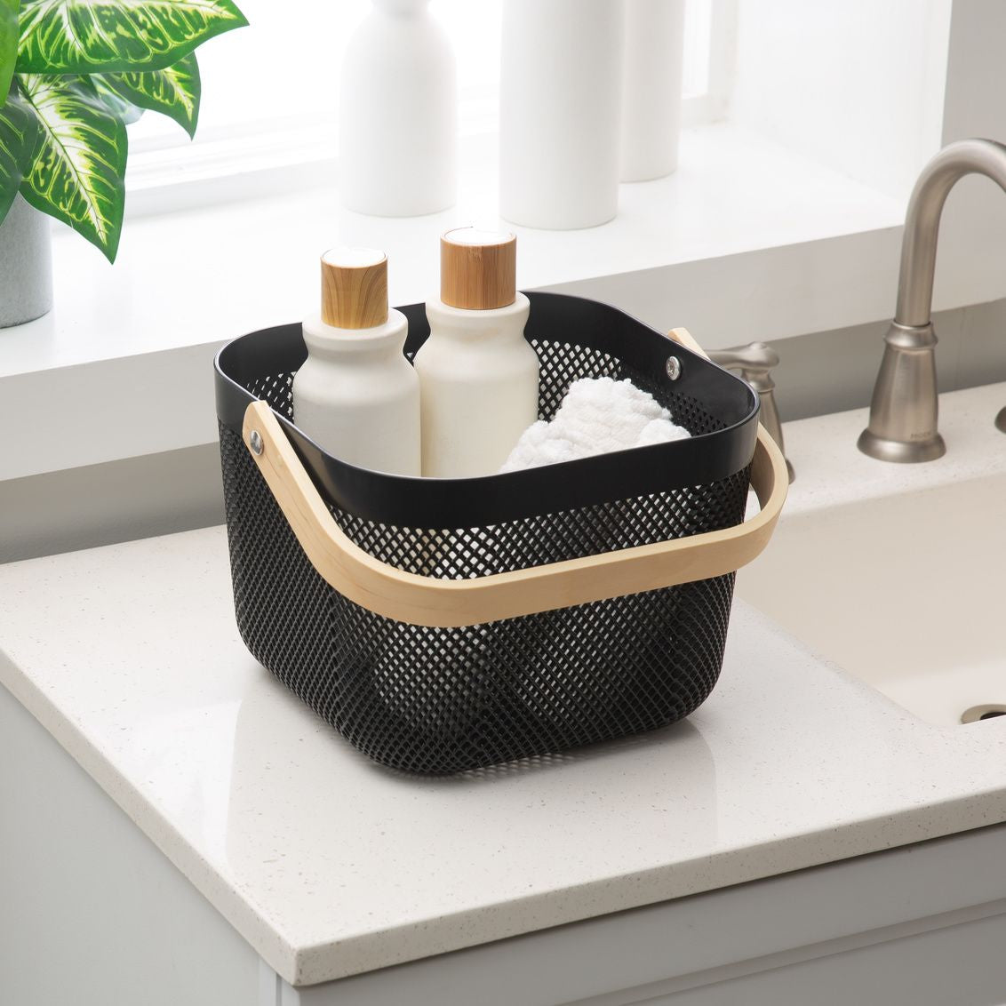 Black mesh storage basket with natural wood handle holding white ceramic bottles and cloth by the kitchen sink