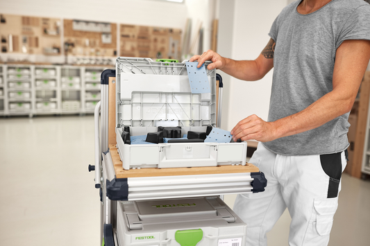 Person organizing blue sanding sheets inside a gray abrasive Systainer on a mobile workbench in a tool accessories store