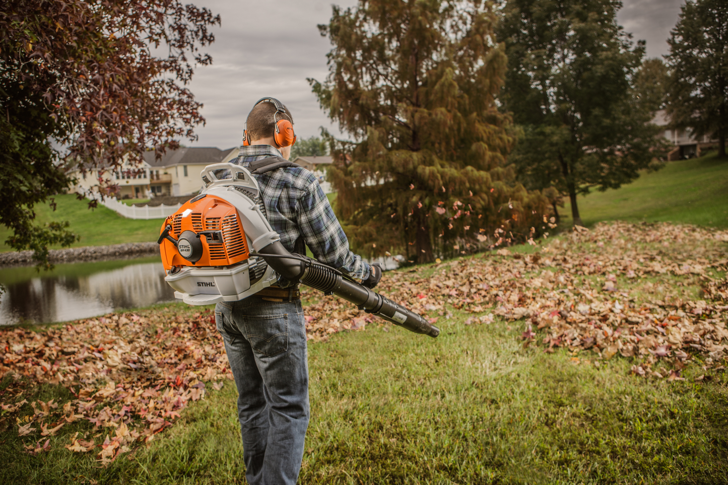 Person using orange and white professional backpack blower to clear leaves from grassy yard near pond and trees