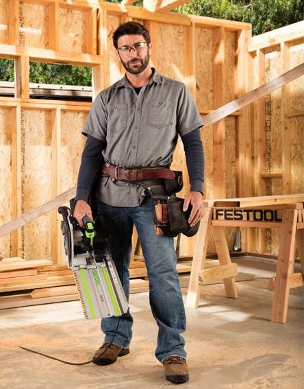 Carpenter holding Festool cordless saw in framed wooden structure, showcasing power tools for home improvement.