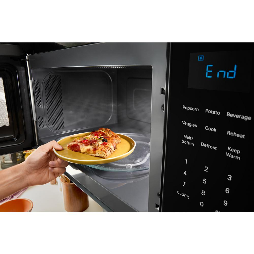 Hand placing a plate with pizza inside a microwave showing digital control panel with sensor cooking options in kitchen appliances collection