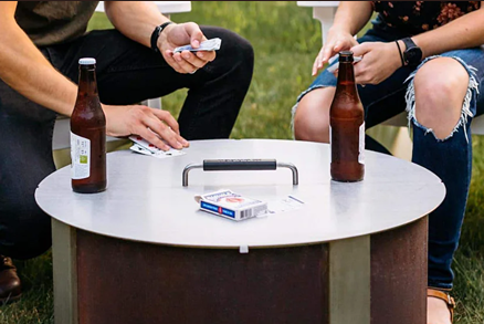 Round metal lid with black handle placed on outdoor fire pit table during casual gathering with drinks and cards