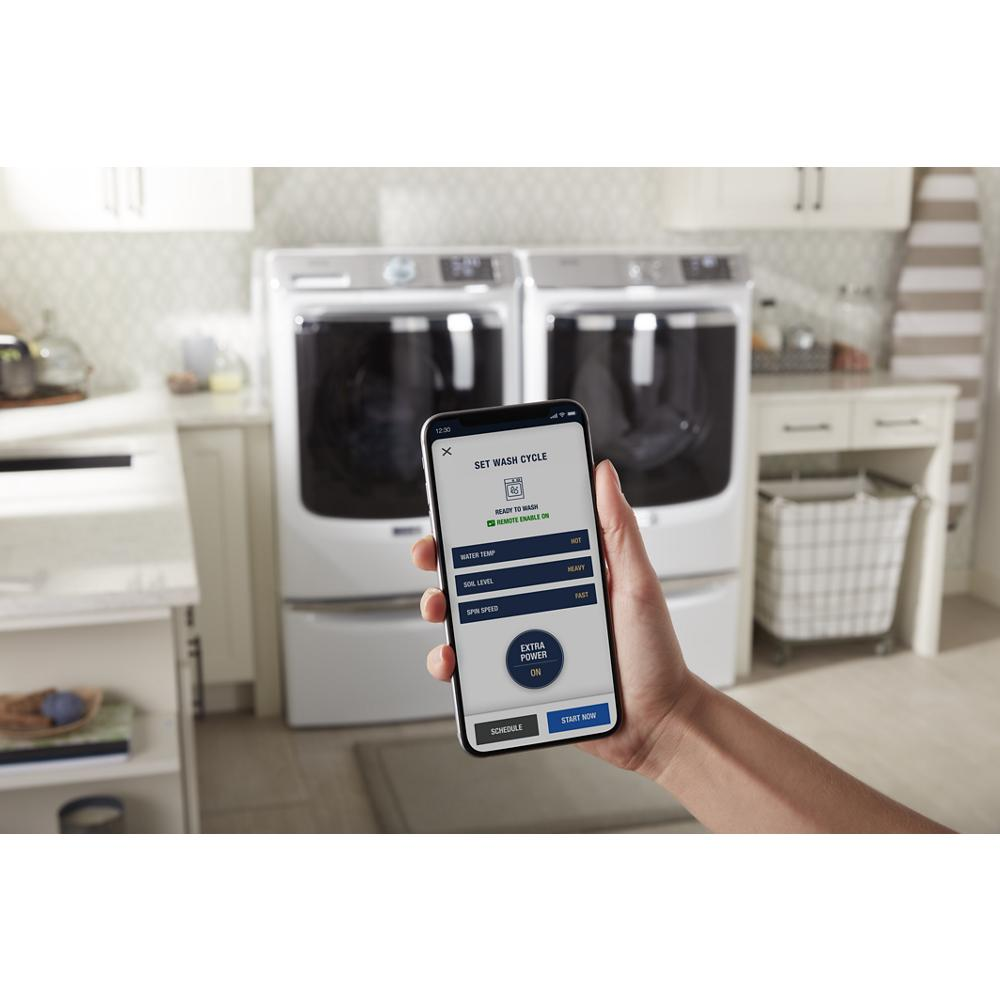 Hand holding smartphone showing laundry cycle settings app with washer and dryer in kitchen laundry room scene