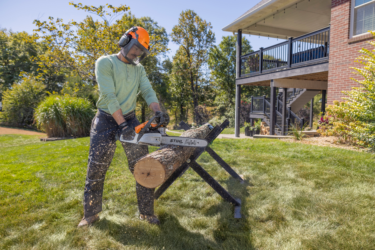 Man wearing safety gear using a STIHL gas-powered chainsaw to cut a log outdoors on a grassy lawn near a house