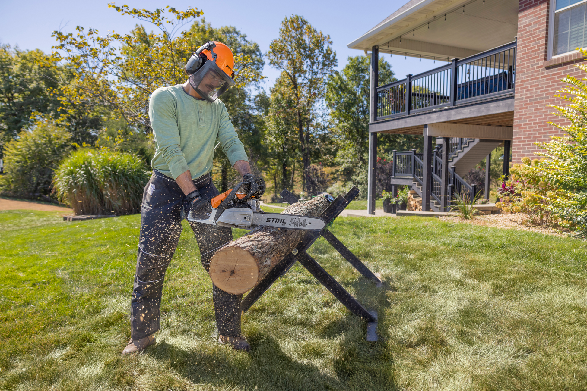 Man wearing safety gear using a STIHL gas-powered chainsaw to cut a log outdoors on a grassy lawn near a house