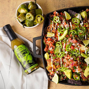 Bottle of Traeger jalapeno lime barbeque sauce next to loaded nachos with jalapenos, lime wedges, and avocado slices