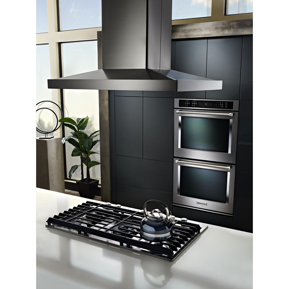 Stainless steel island-mount canopy hood above gas cooktop with dual ovens in modern kitchen setting