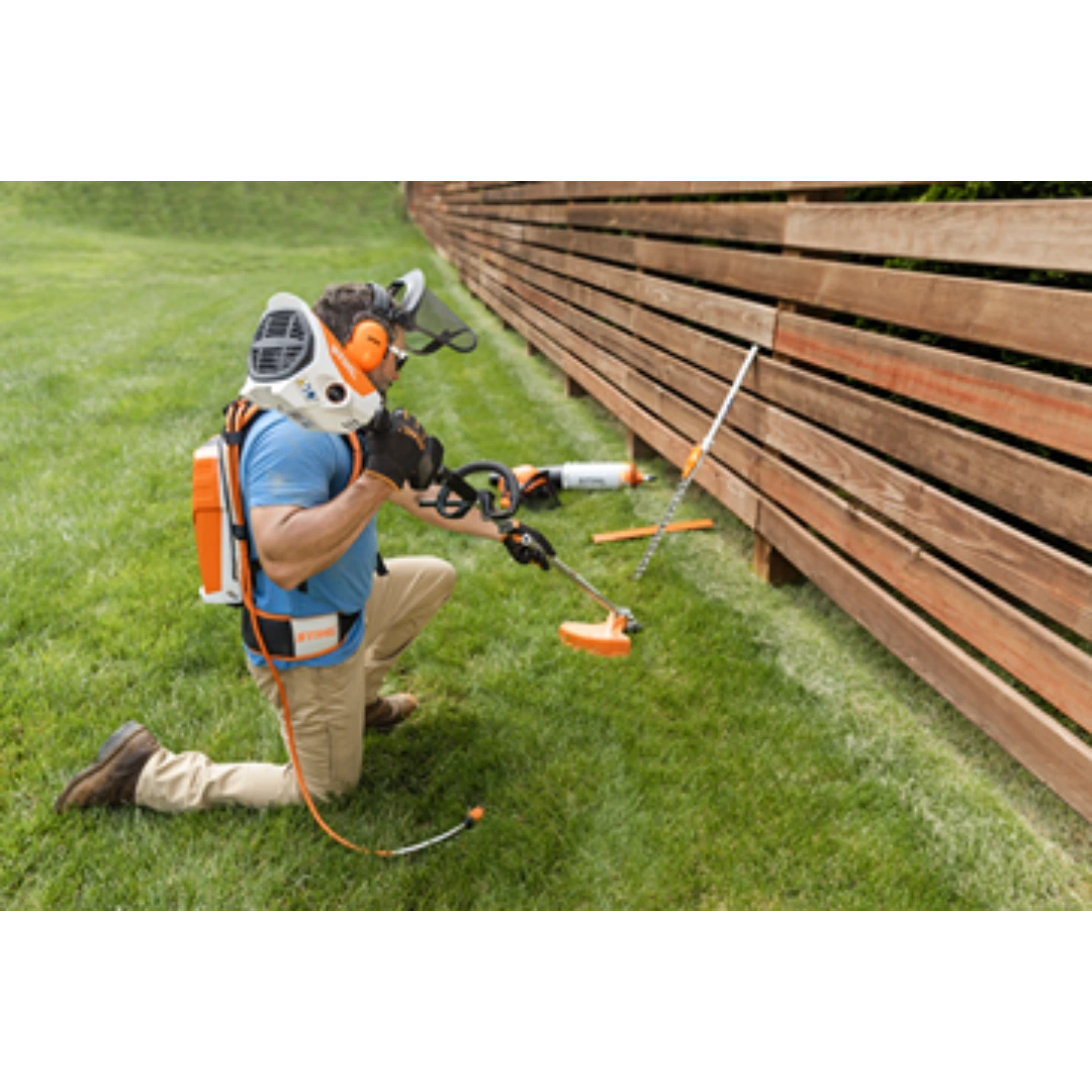 Person wearing protective gear using a STIHL cordless trimmer to edge grass along a wooden fence in outdoor power equipment setting