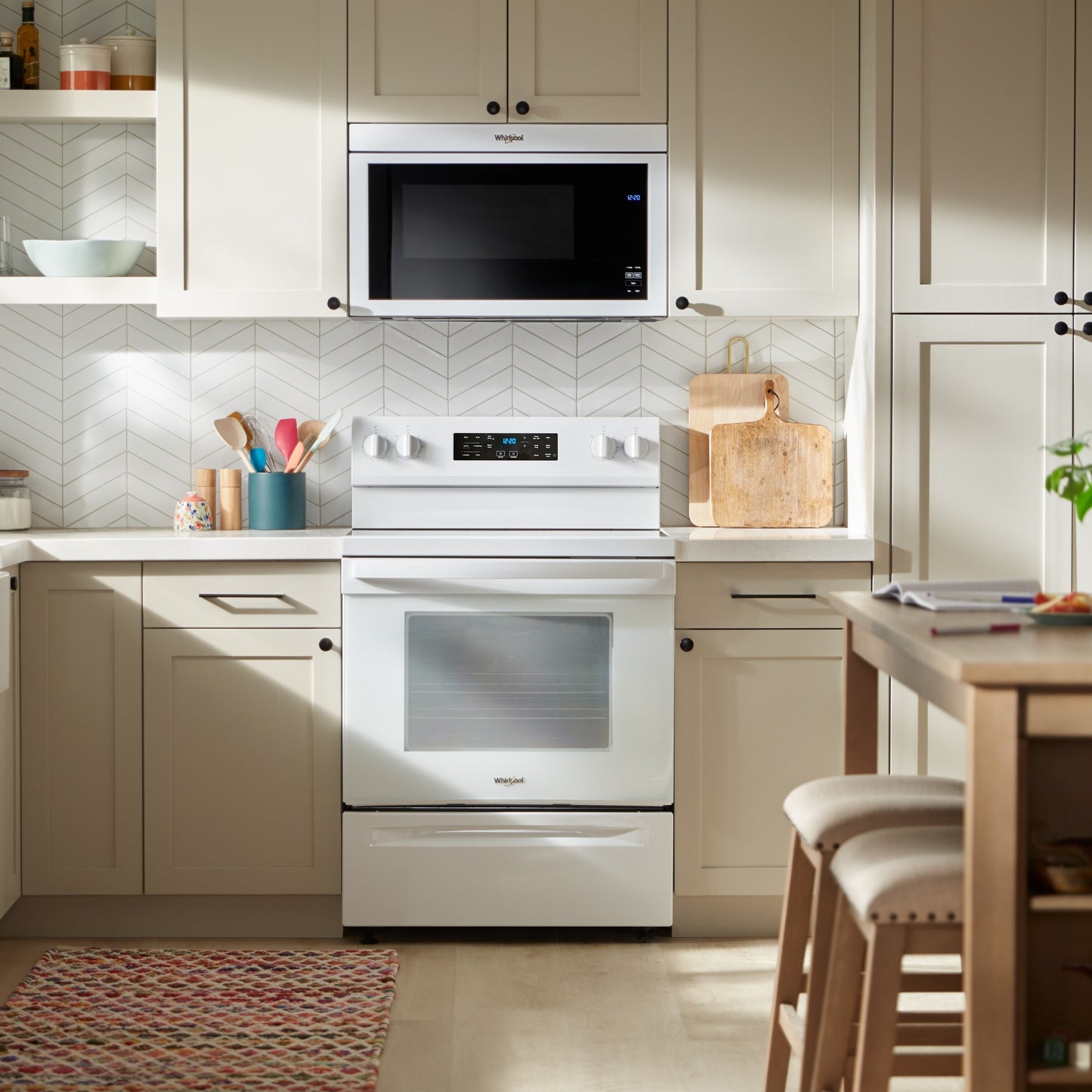 Modern white electric range and microwave built into beige kitchen cabinetry with countertop accessories and patterned rug
