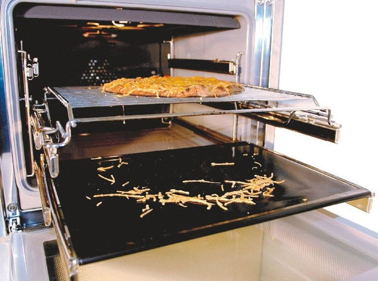 Black reusable oven liner protector placed on lower rack catching melted cheese for easy cleanup in home kitchens