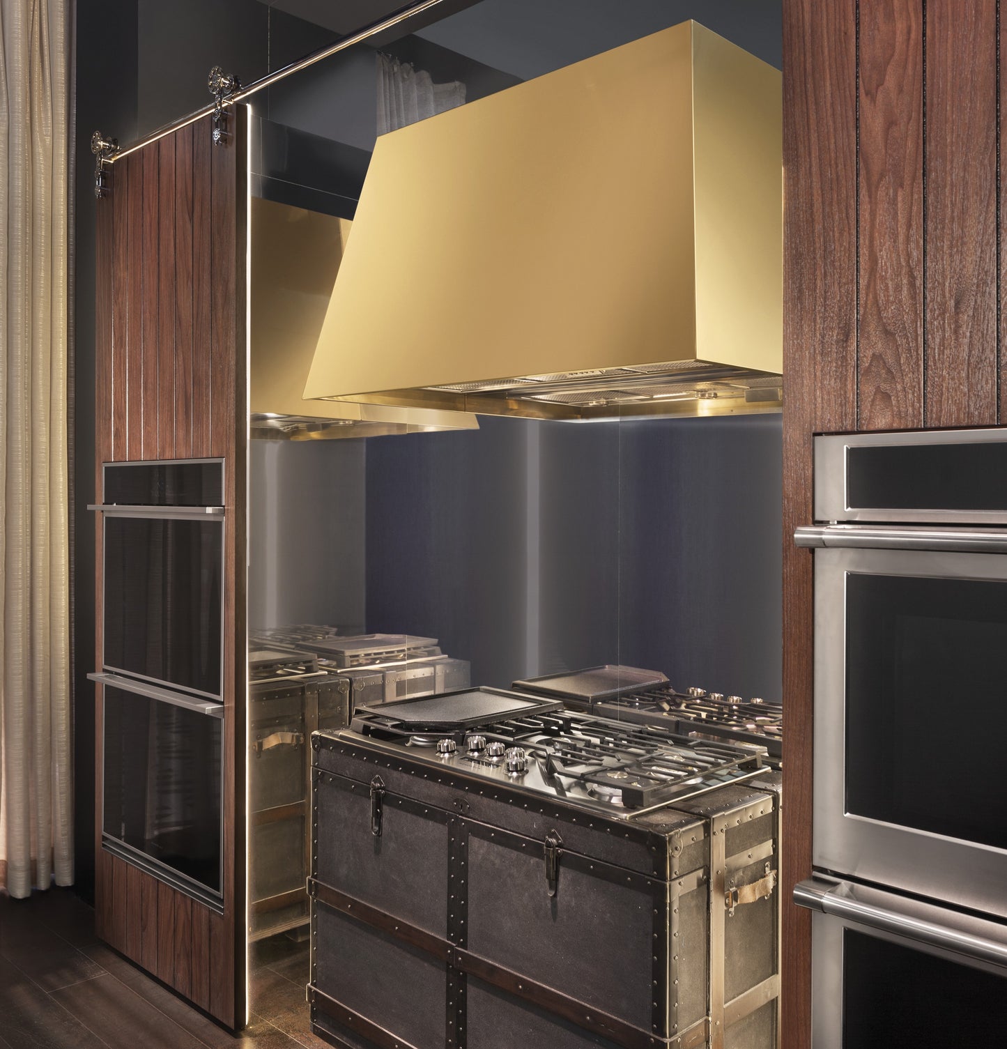 Contemporary kitchen with wood paneled double wall ovens beside a large brass range hood and vintage-style stove trunk unit