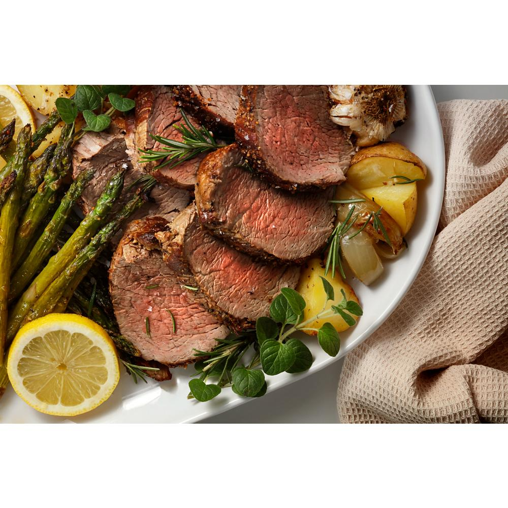 Close-up of sliced roast beef with grilled asparagus, roasted potatoes, lemon slice, and fresh herbs on white plate