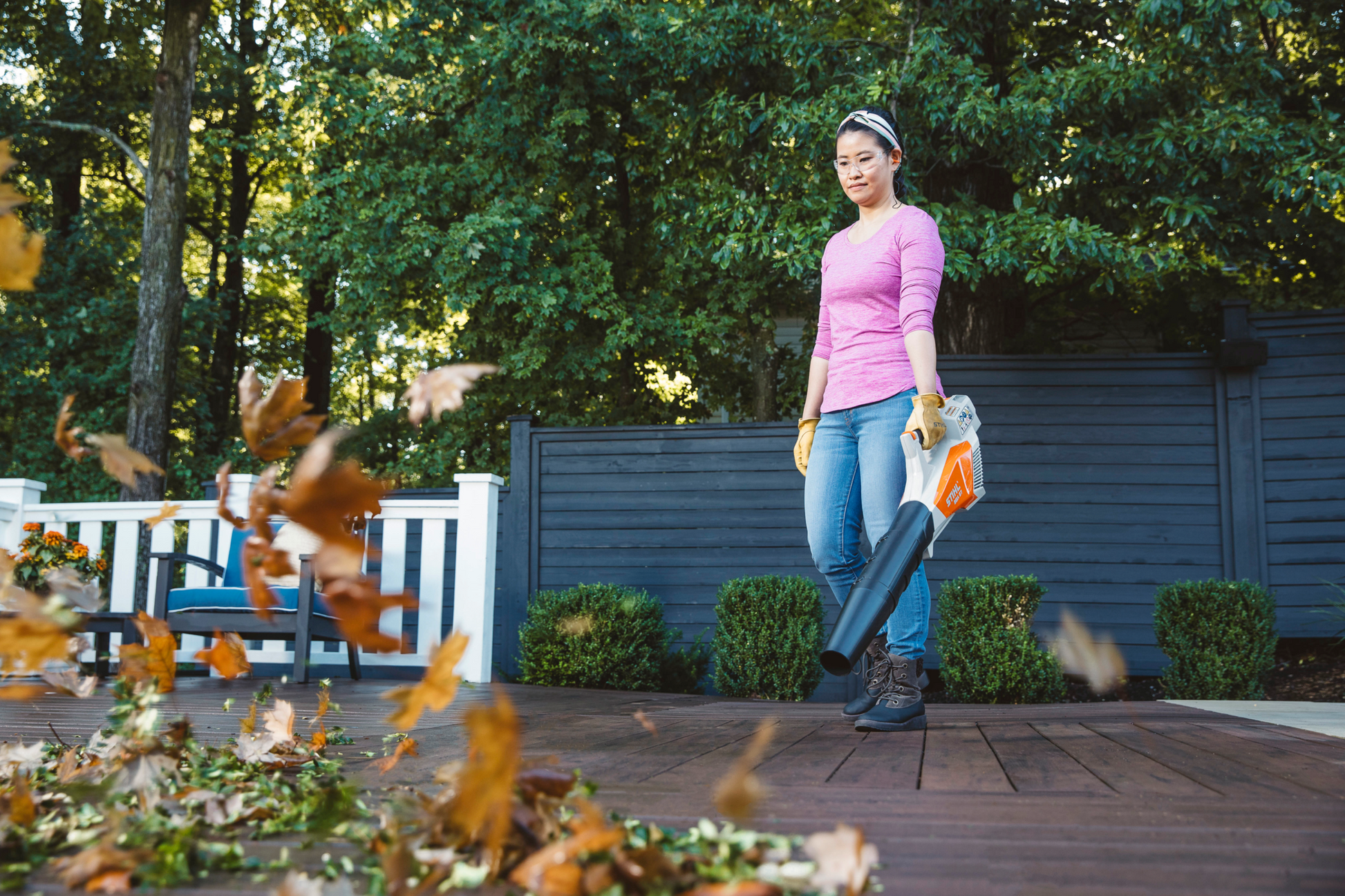 Woman wearing gloves using a handheld blower to clear leaves from a wooden deck in a backyard setting