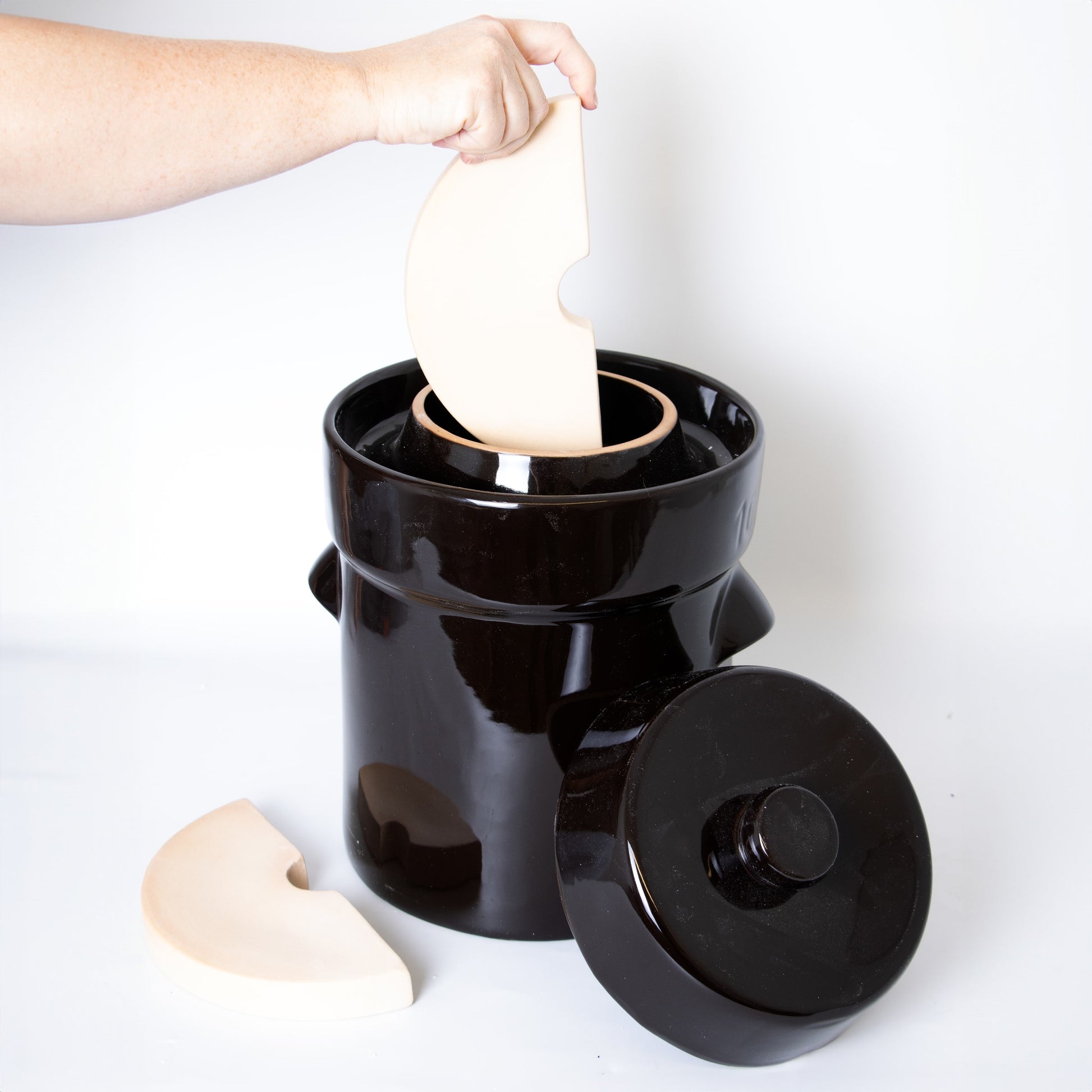 Hand inserting ceramic weight into large dark crock with matching lid on white background in kitchenware setting