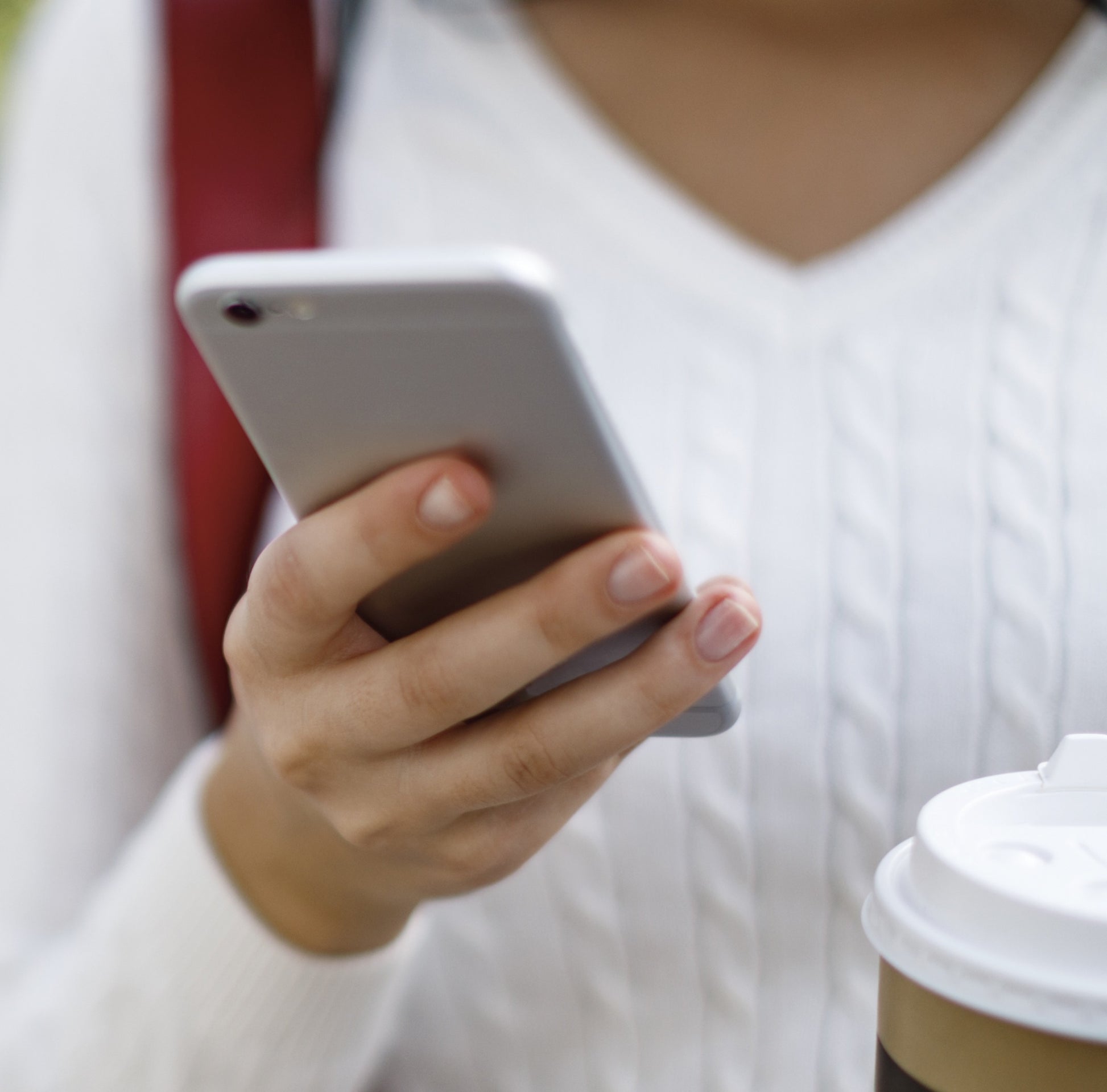 Close-up of person holding smartphone and coffee cup wearing white sweater, suggesting casual home or kitchen setting.