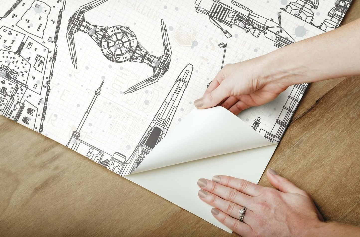 Hands peeling back white and black Star Wars blueprint peel and stick wallpaper on a wood surface