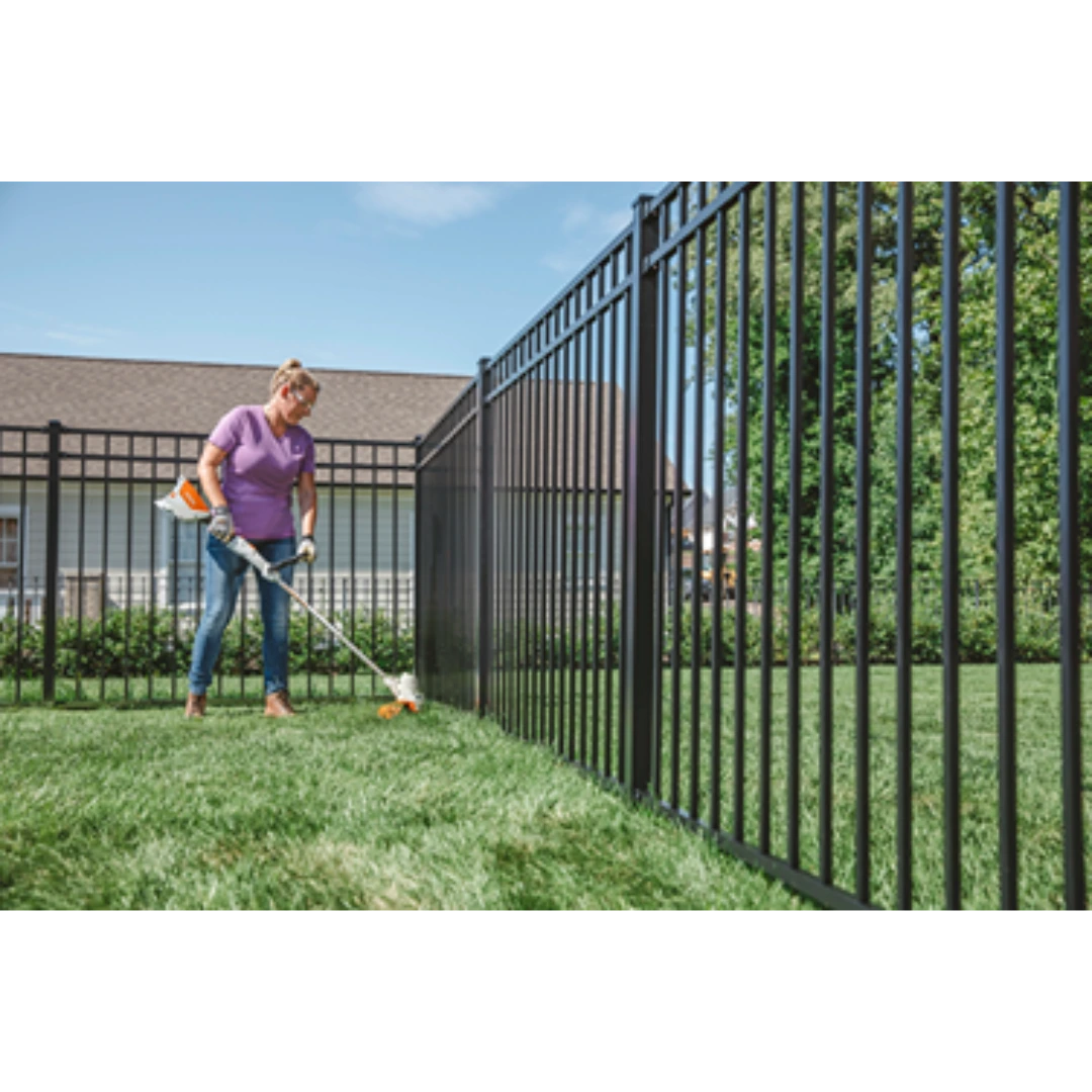 Person using battery-powered cordless trimmer on grass along black metal fence for outdoor home improvement tasks