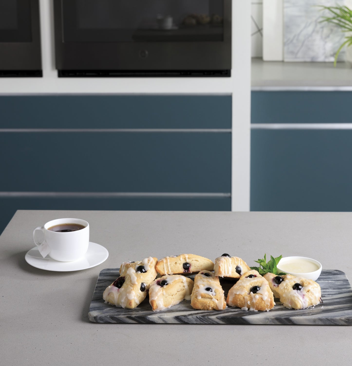 Blueberry scones on a marble board with white sauce and cup of black coffee on kitchen island near wall ovens