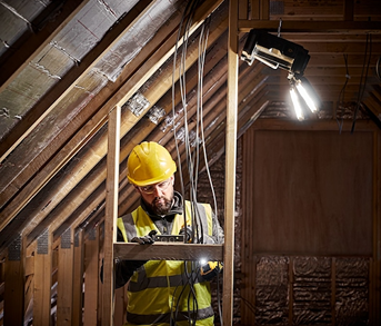 Construction worker wearing yellow hard hat using adjustable LED work light for attic insulation in home improvement setting