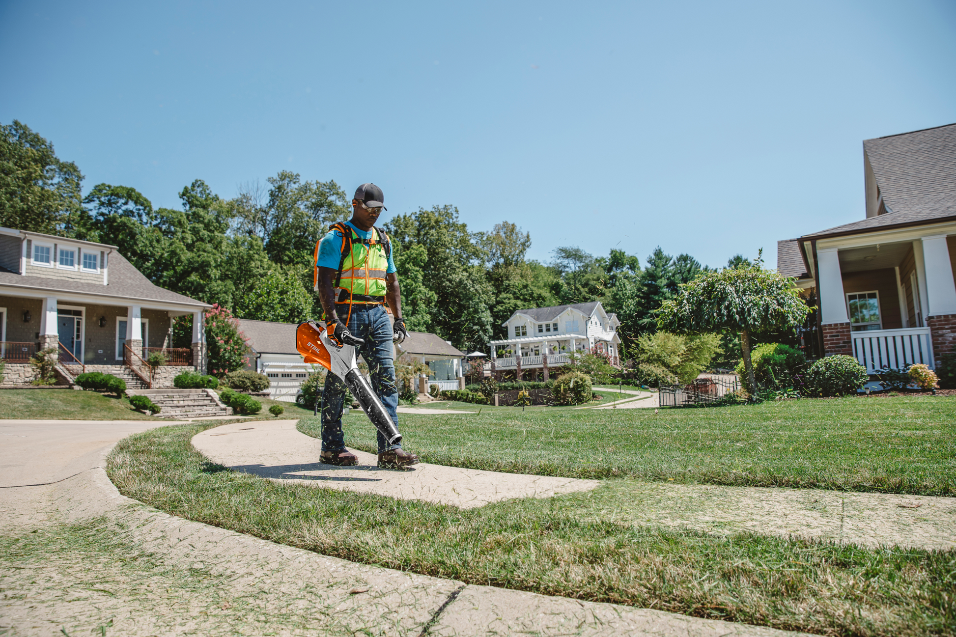 Worker wearing safety vest using orange handheld battery blower to clear grass from residential curved concrete walkway edge