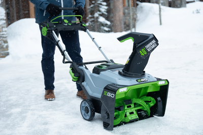 Person operating green and gray cordless snow blower with steel auger clearing snow on driveway for home improvement use