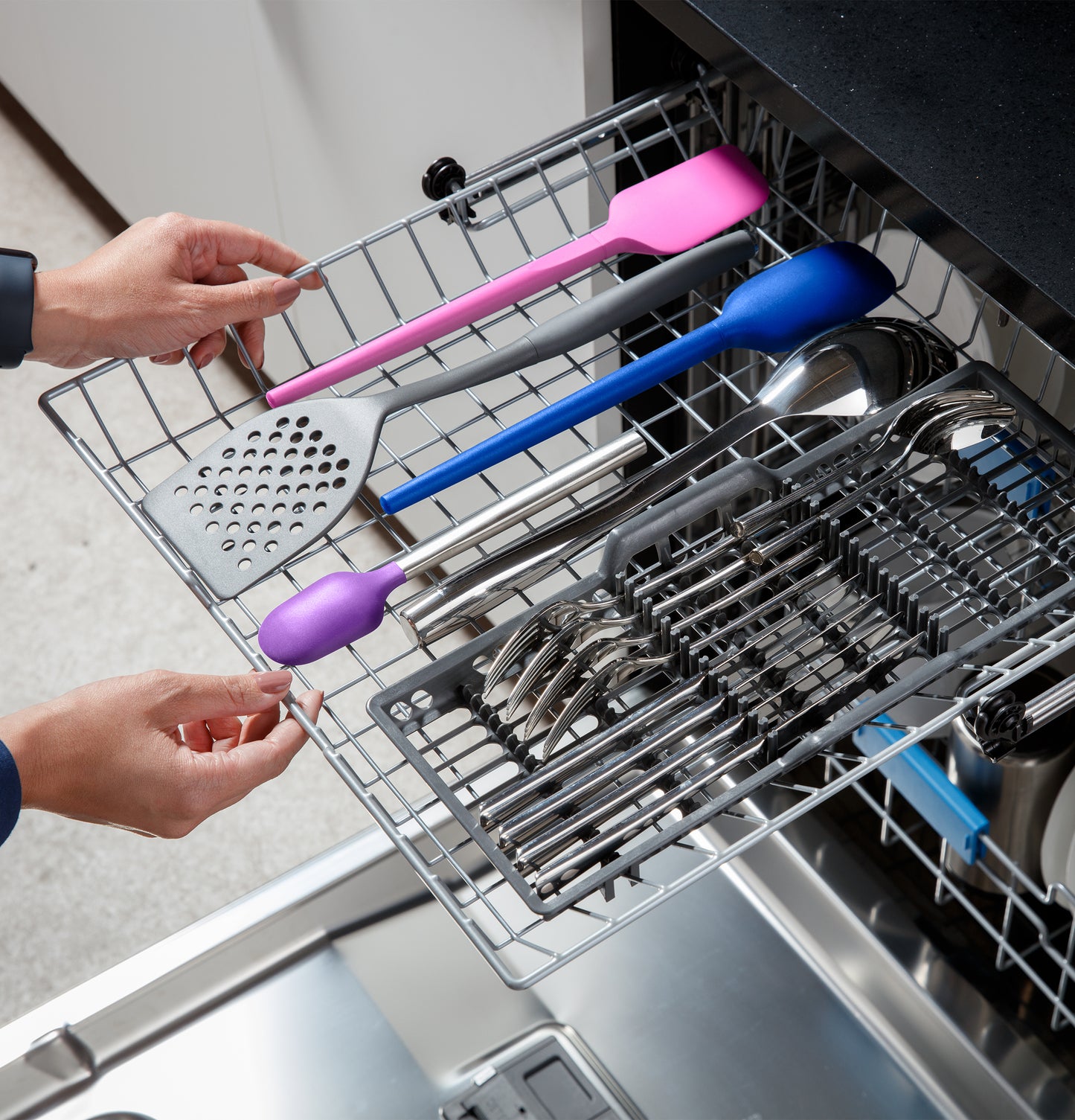 Top rack of stainless steel dishwasher loaded with colorful kitchen utensils and cutlery in modern appliance setting