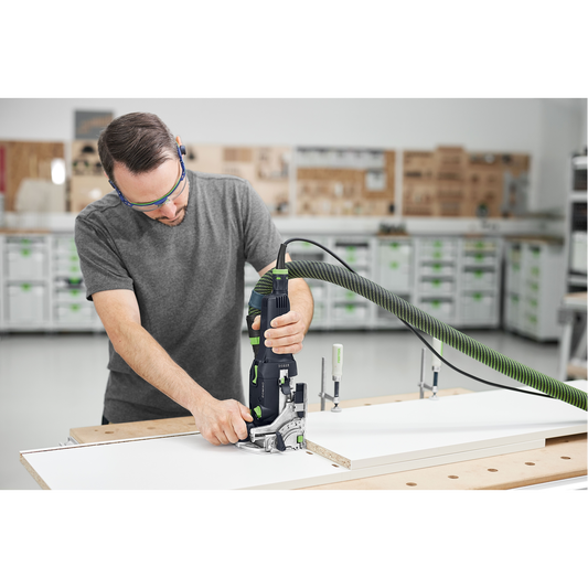 Man using a Festool DOMINO joiner with dust extraction hose to cut precise joints on a white wood board in a woodworking workshop surrounded by power tools and storage cabinets.