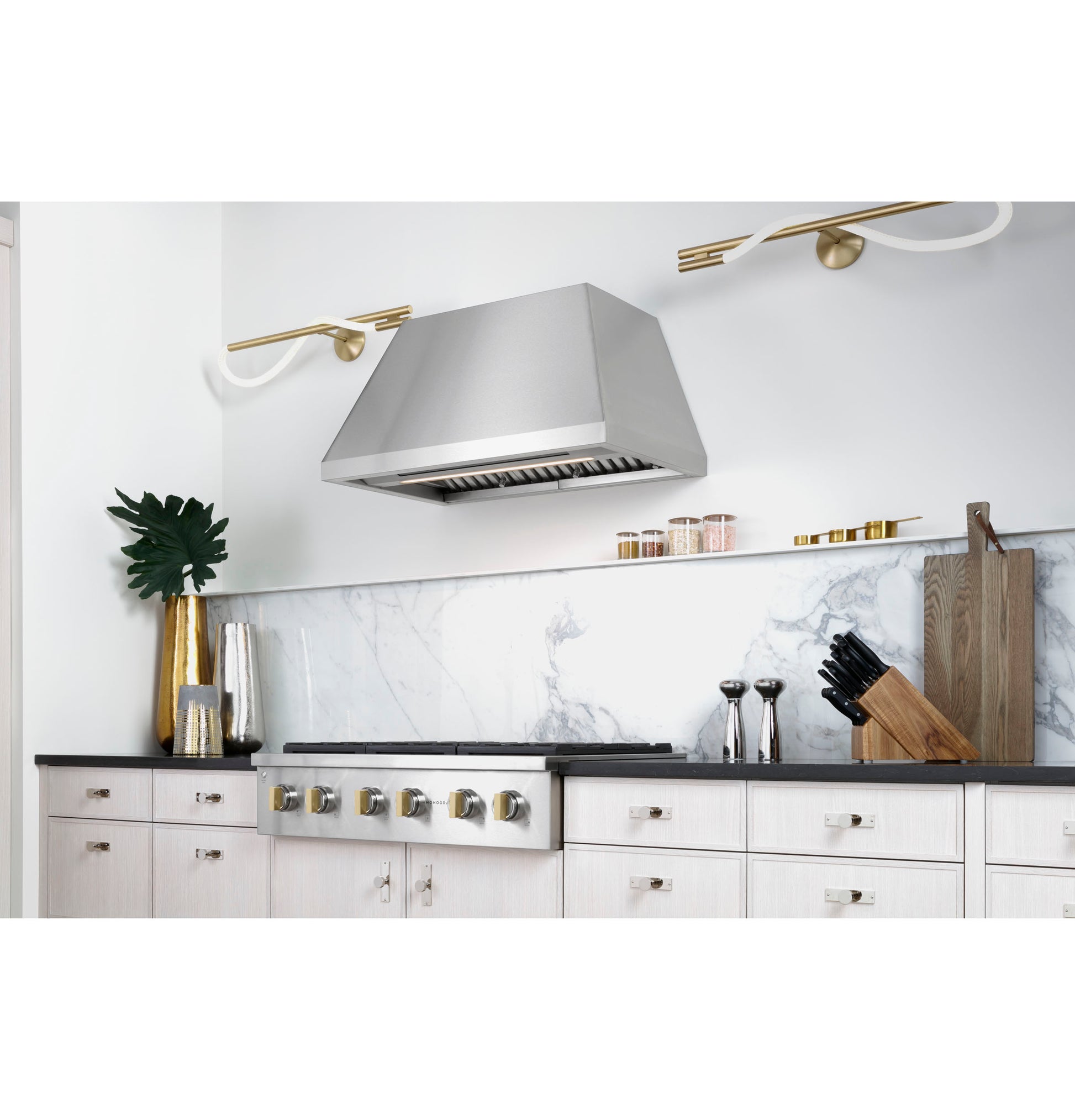 Modern kitchen cooktop area featuring a stainless steel vent hood, marble backsplash, and white cabinetry with black countertop