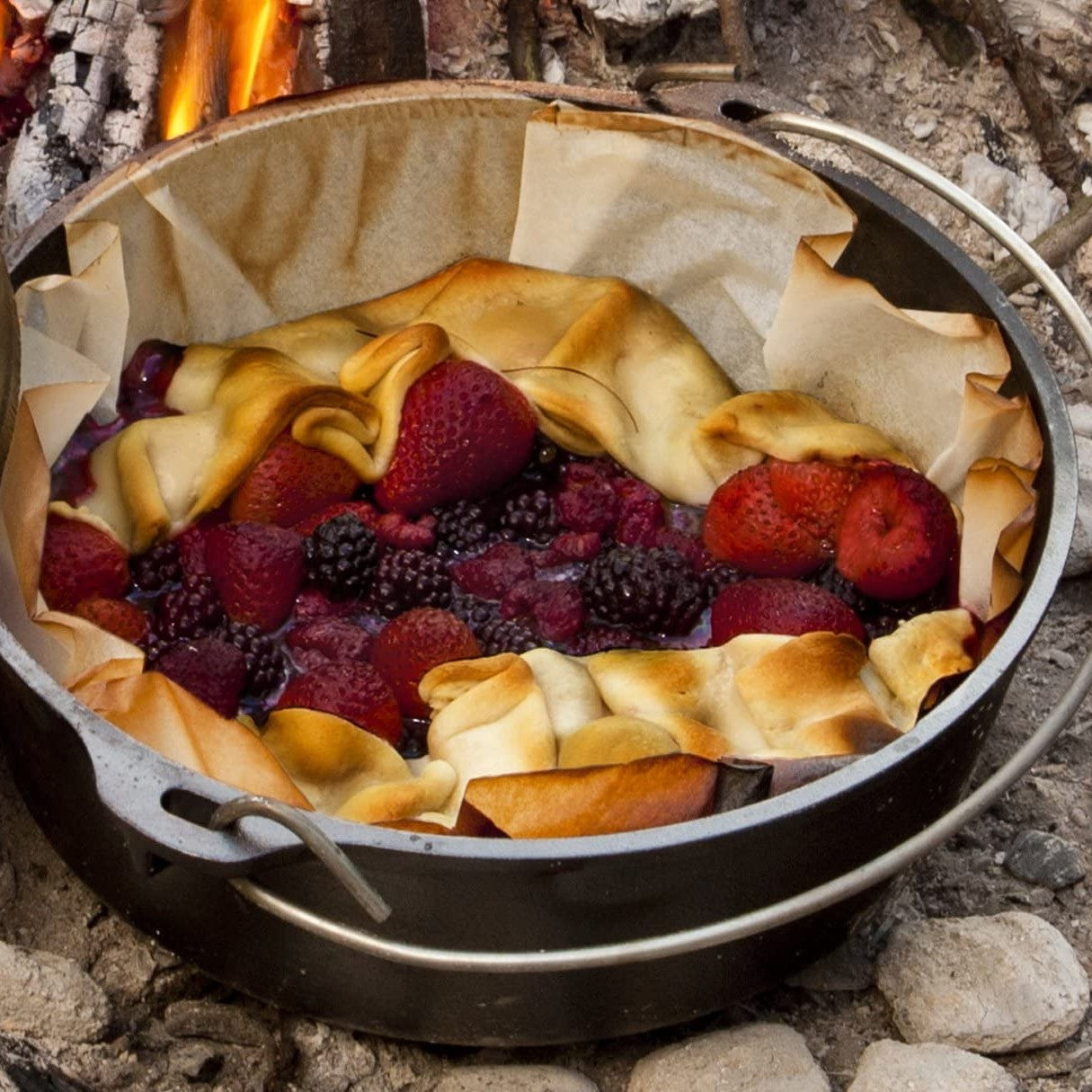 Dutch oven lined with parchment paper baking a mixed berry dessert over campfire in outdoor kitchenware setup