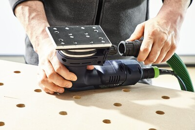 Person attaching green dust extraction hose to blue cordless orbital sander on wooden workbench for home improvement tasks