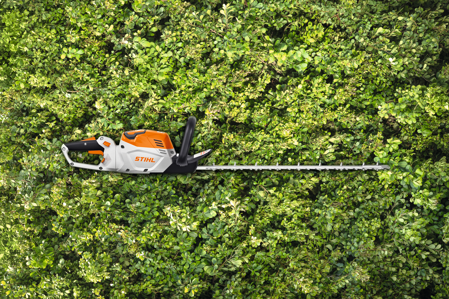 Electric hedge trimmer with orange and white body and long blade resting on dense green hedge for home improvement tasks