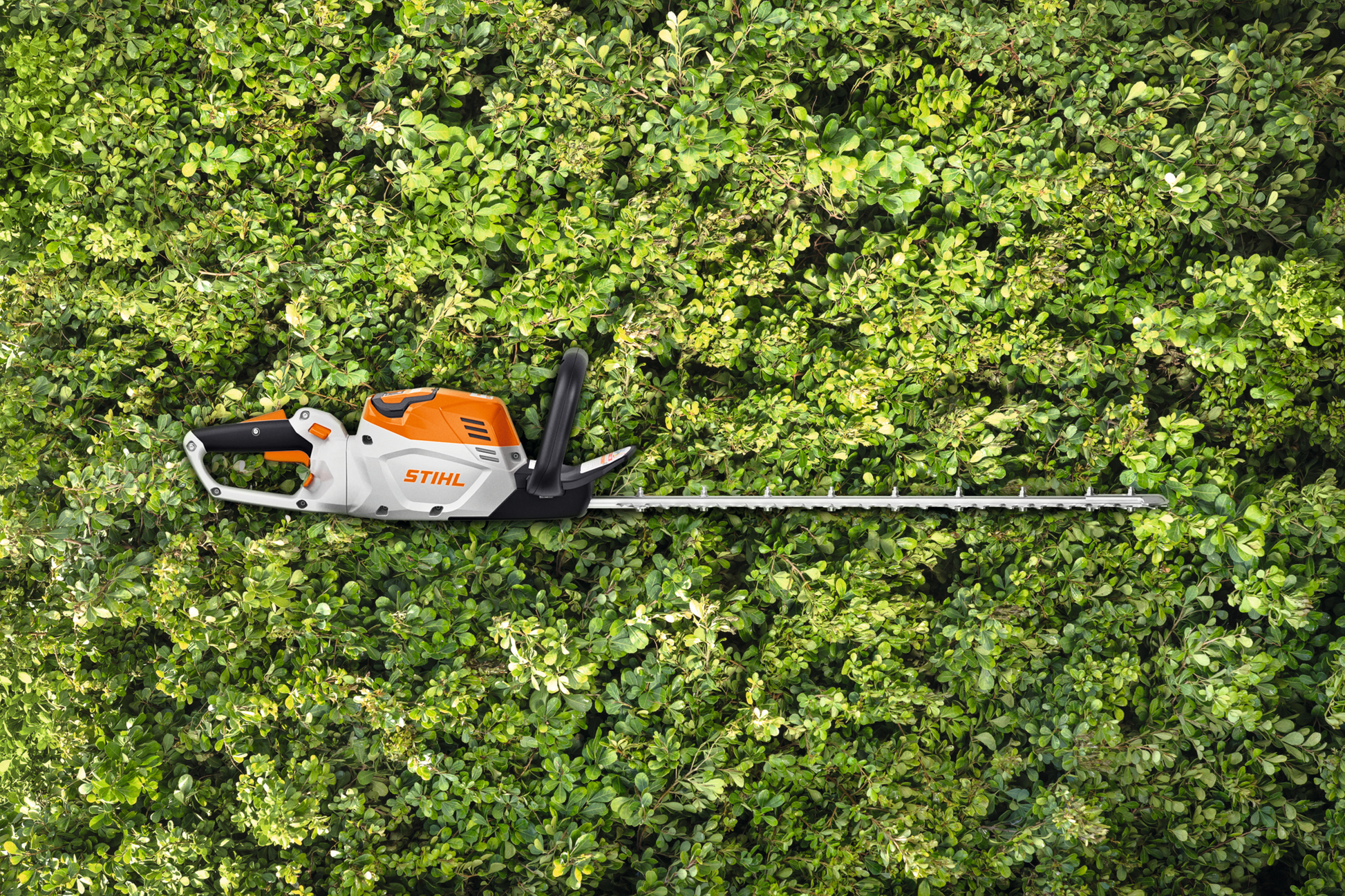 Electric hedge trimmer with orange and white body and long blade resting on dense green hedge for home improvement tasks