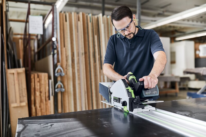 Man wearing safety glasses using a power tool with guide rail for woodworking in a workshop setting