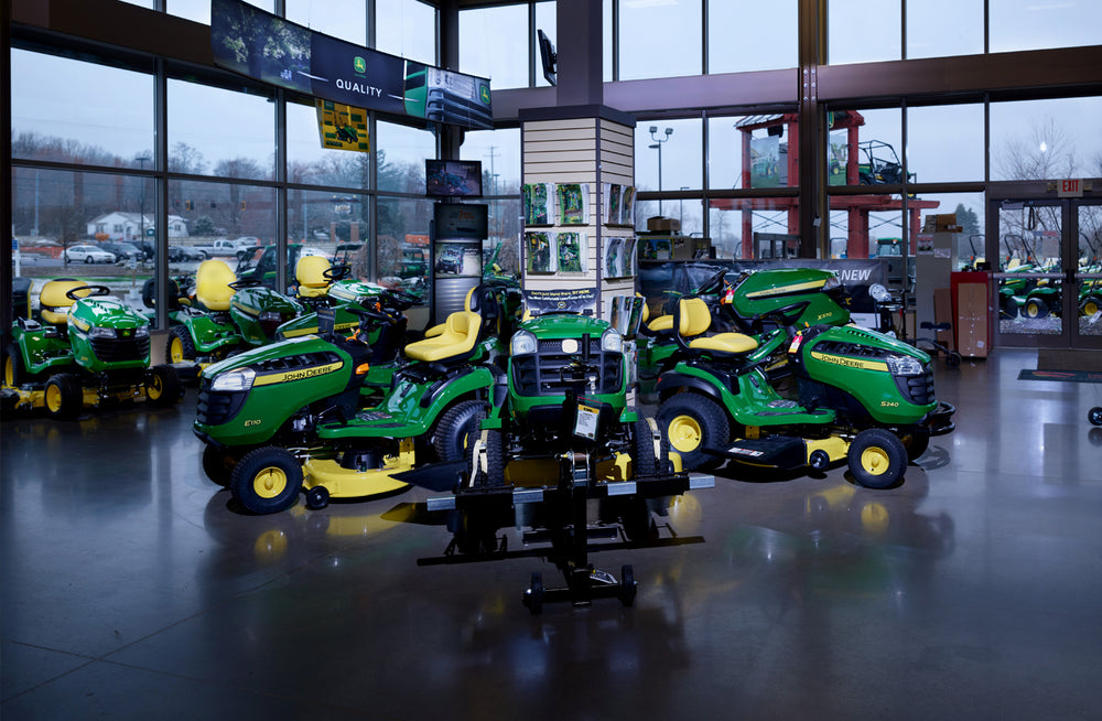 Indoor showroom featuring multiple green and yellow lawn tractors and attachments displayed under large windows with outdoor views visible.