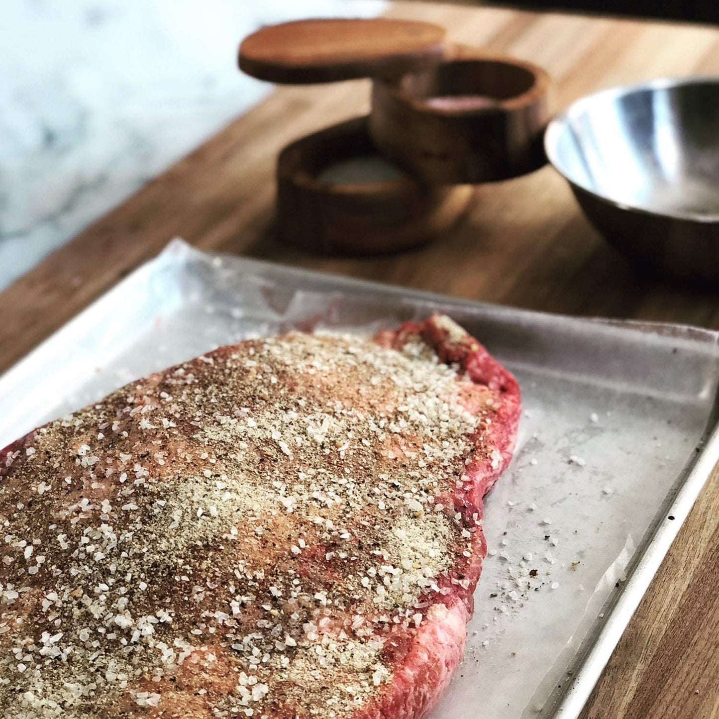 Round wooden salt cellar made of Acacia wood with hinged lid next to seasoned raw meat on cutting board