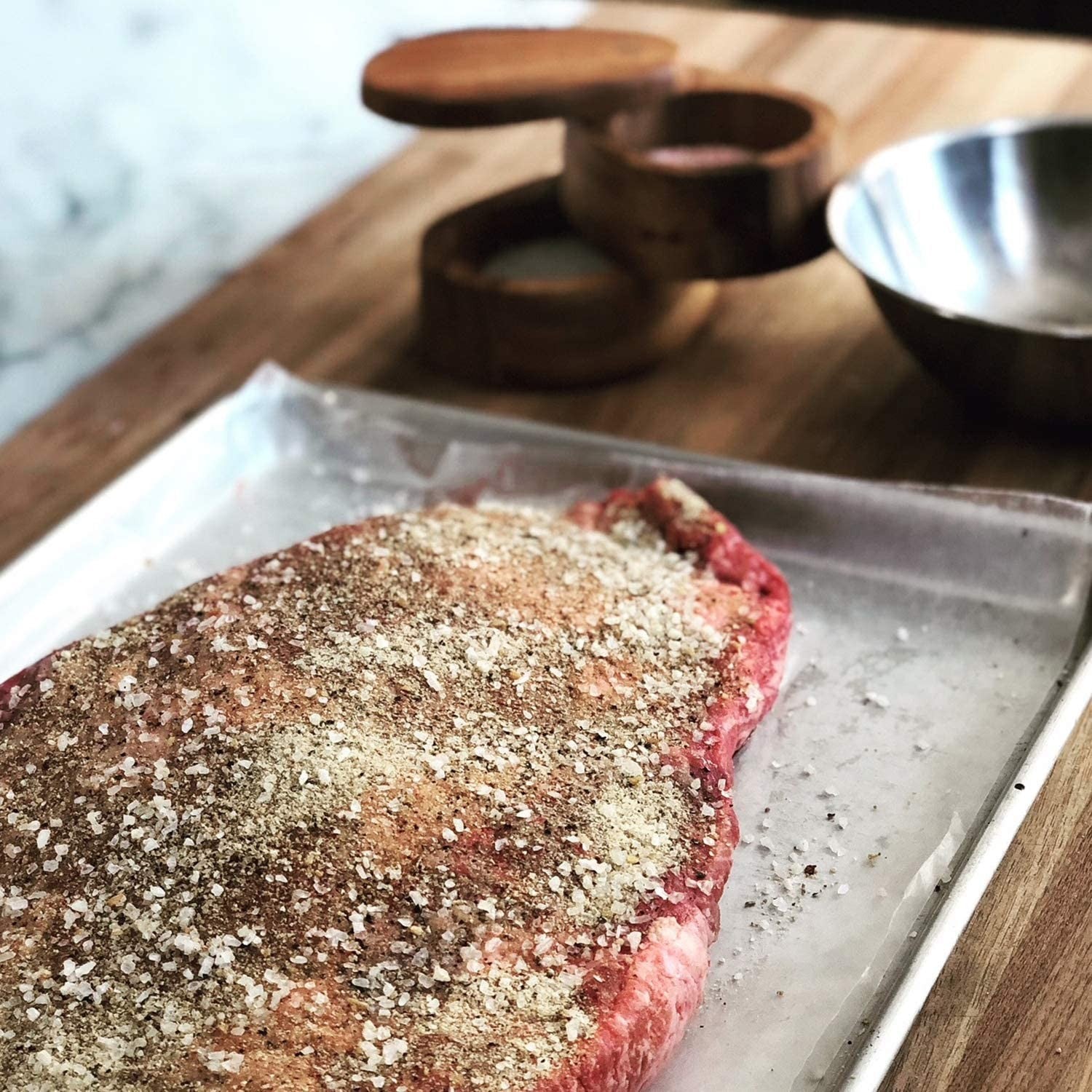 Round wooden salt cellar made of Acacia wood with hinged lid next to seasoned raw meat on cutting board