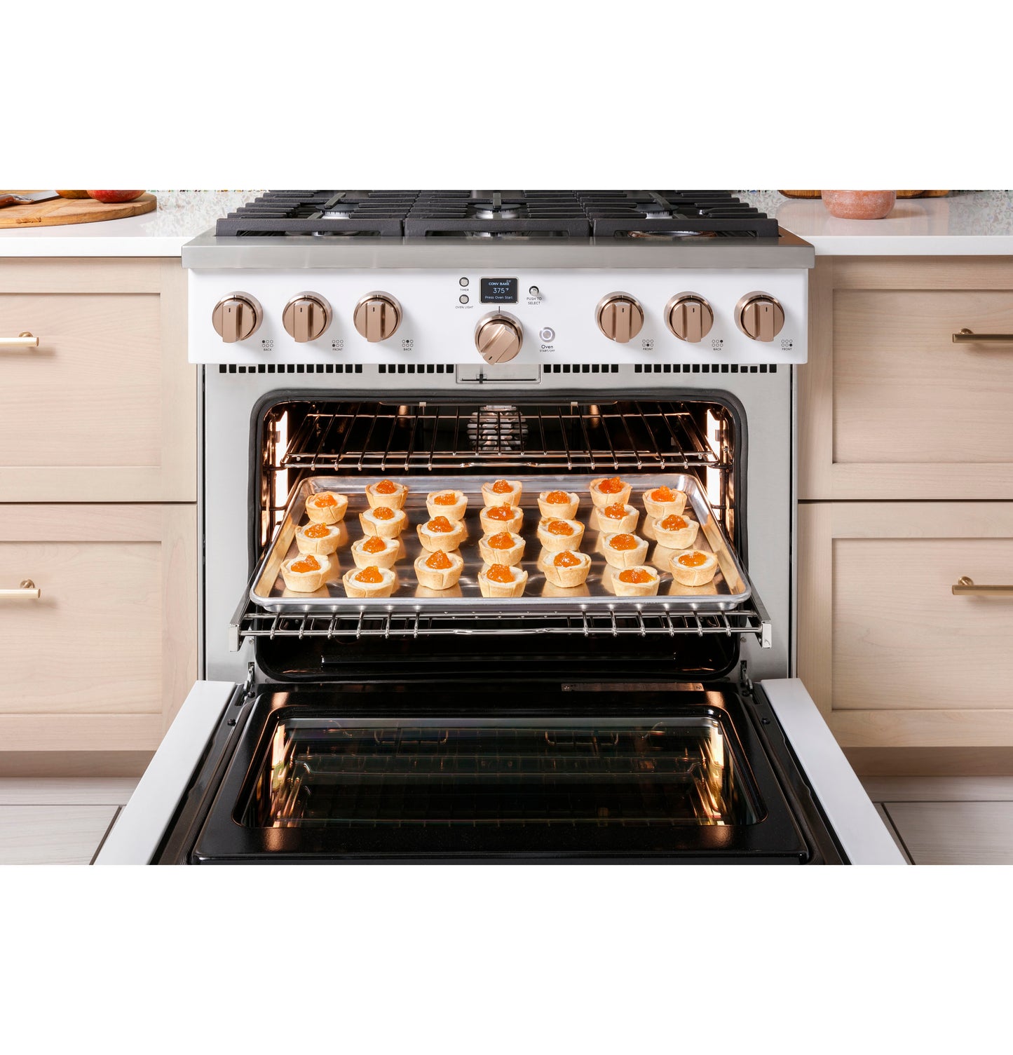 White gas range with six burners and open oven door showing a tray of pastries baking in modern kitchen setting