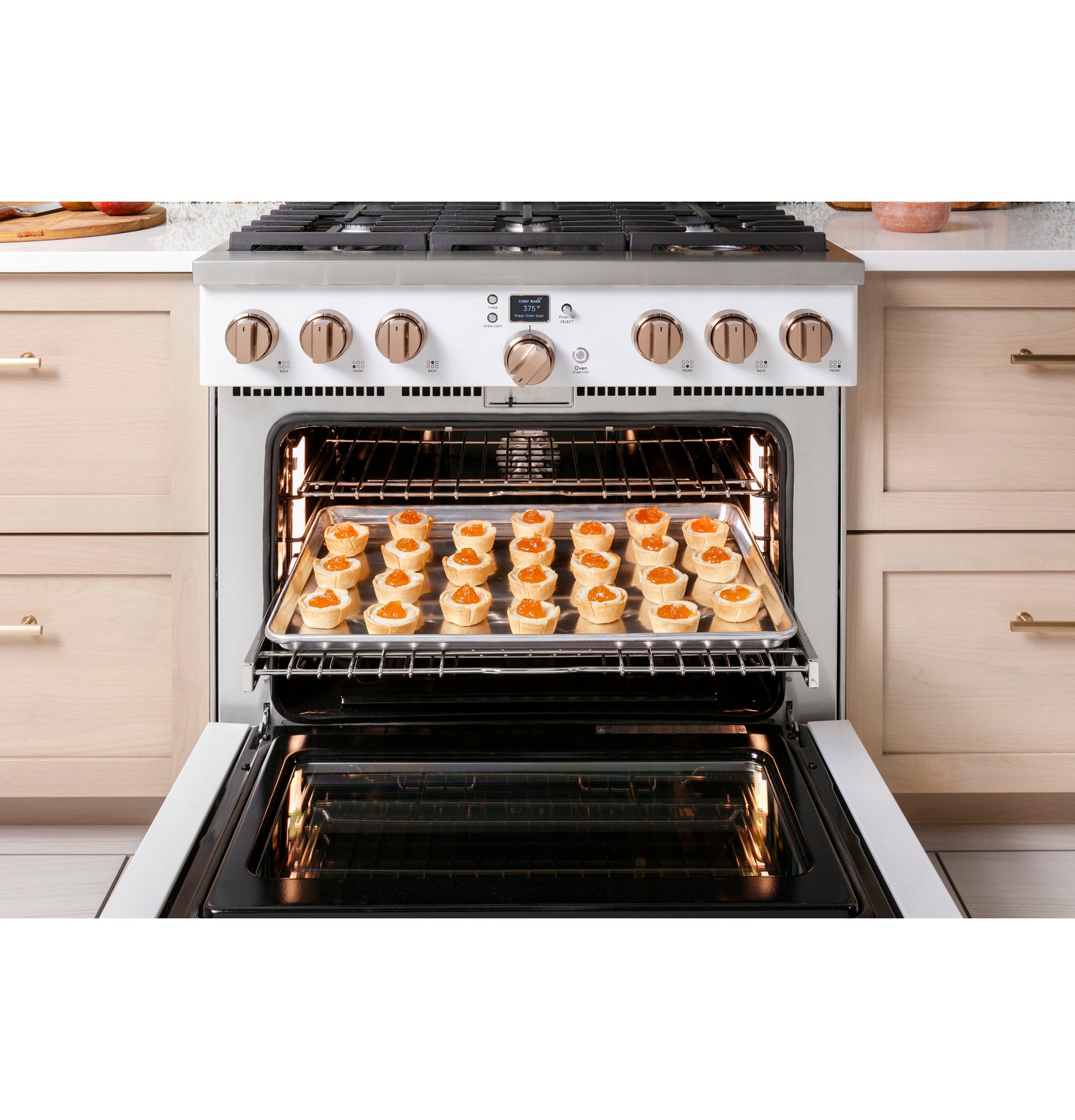 White gas range with six burners and open oven door showing a tray of pastries baking in modern kitchen setting
