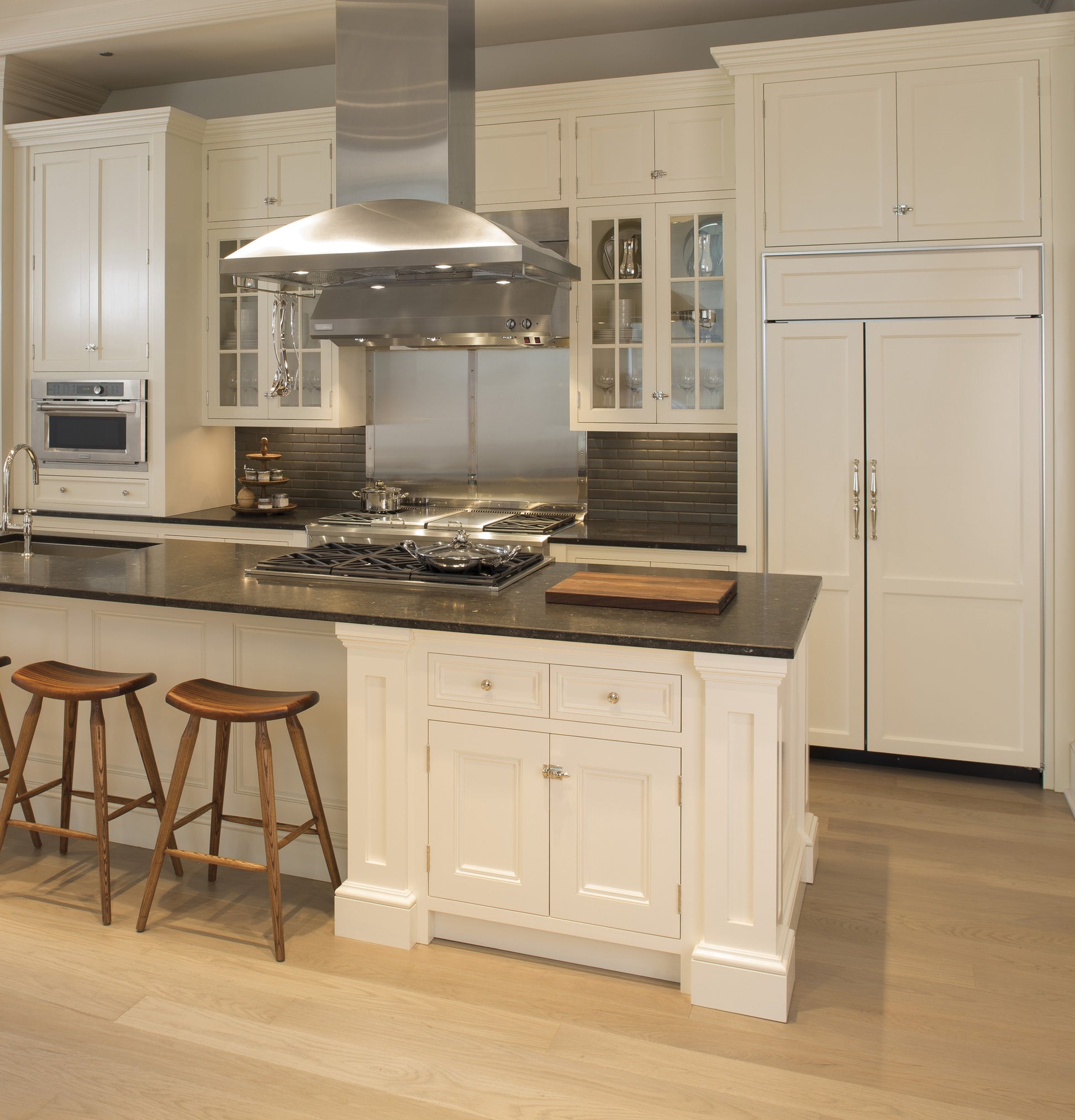 Stainless steel island hood with chimney vent over cooktop in white kitchen with wood stools and cabinetry